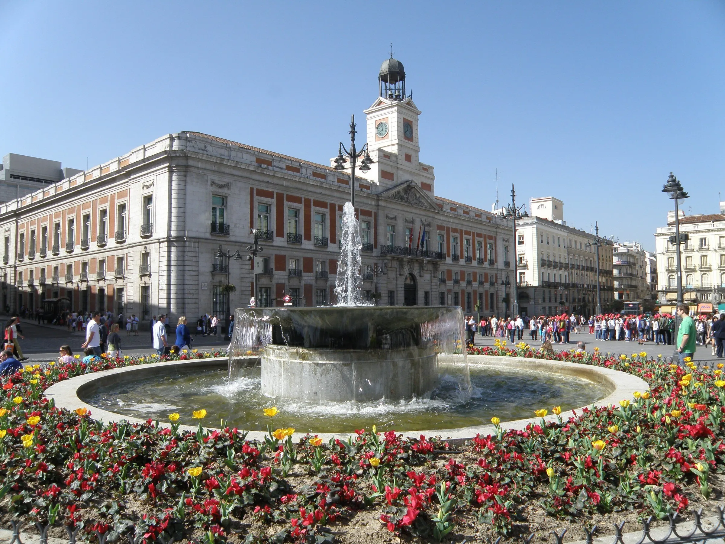 Casa de Correos and its clock in the Puerta del Sol, with a fountain, flowers and pedestrians in front.