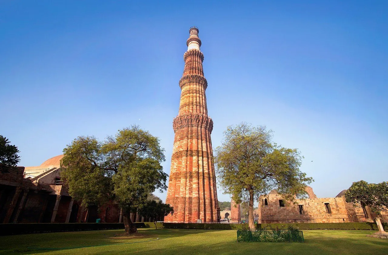 Torre de arenisca roja del Qutub Minar, en Delhi, con franjas de inscripciones, entre árboles y ruinas, bajo cielo azul.