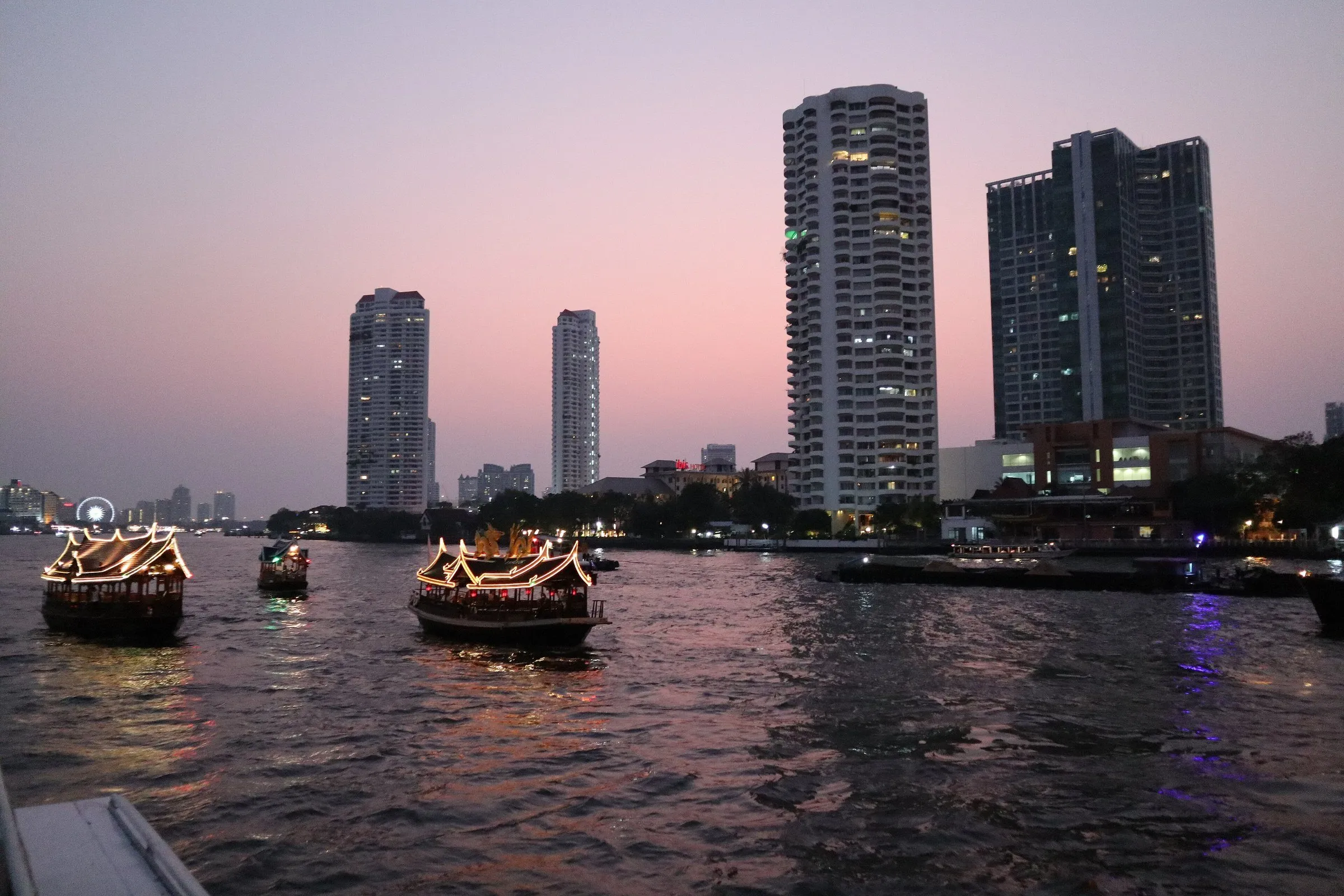 Barcos de passeio com contornos iluminados no rio Chao Phraya, ao entardecer, com prédios altos na margem e céu rosado.
