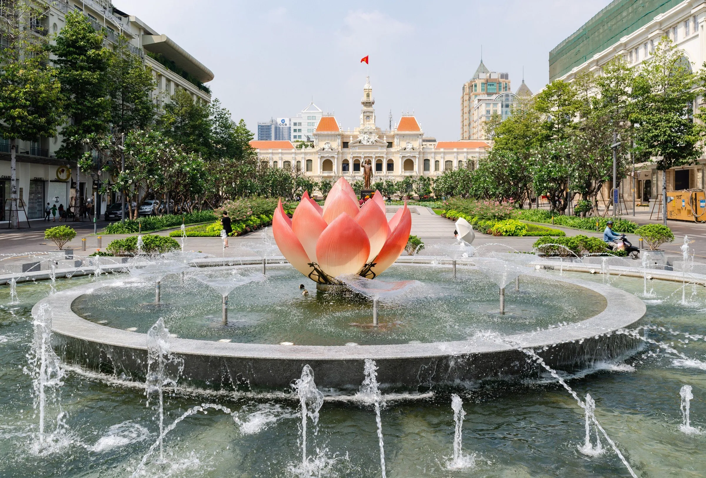 Fonte com lótus rosa no eixo da rua Nguyễn Huệ, com jardins e o edifício do Comitê Popular ao fundo.