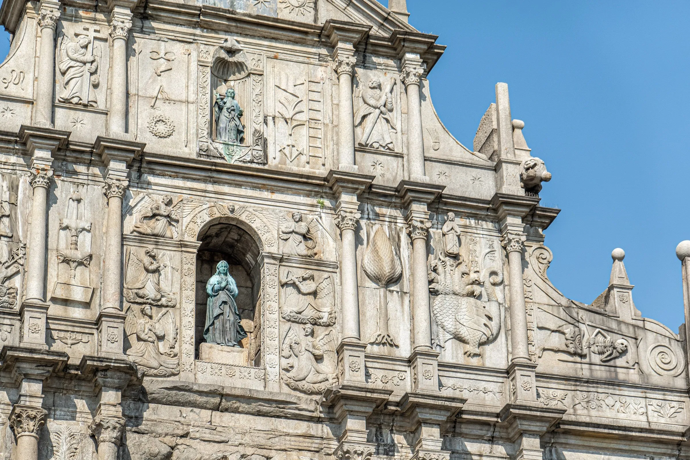 Detalle de la fachada tallada de las Ruinas de San Pablo, con estatuas de bronce, ángeles en relieve y cielo azul al fondo.