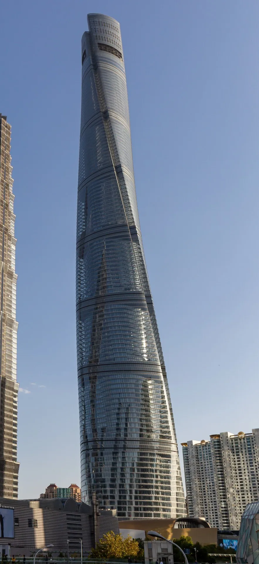 Shanghai Tower in a vertical shot, with a twisting glass facade, blue sky and buildings at its base.