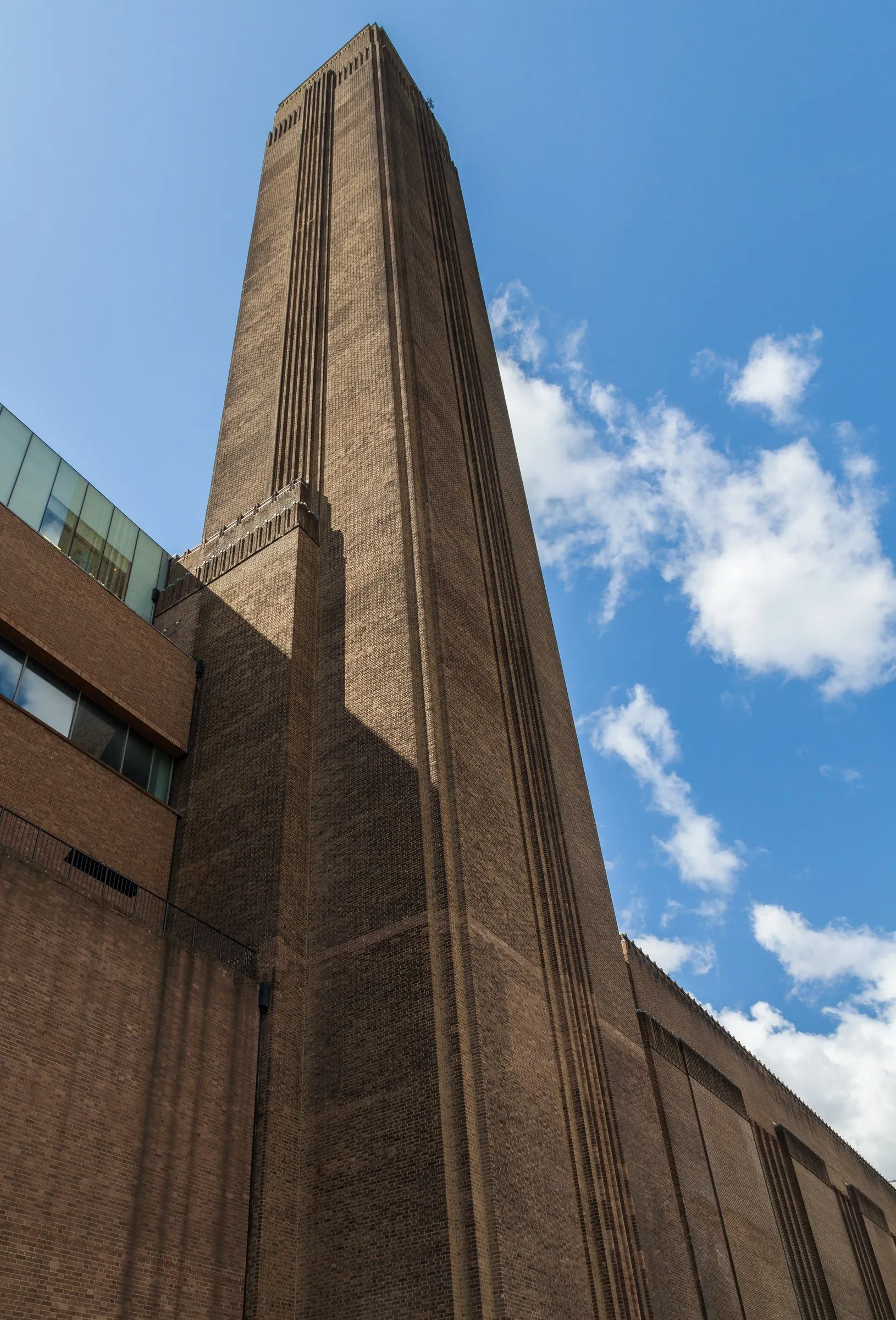 Brick chimney of Tate Modern, seen from below against a blue sky with clouds, next to the brick building.