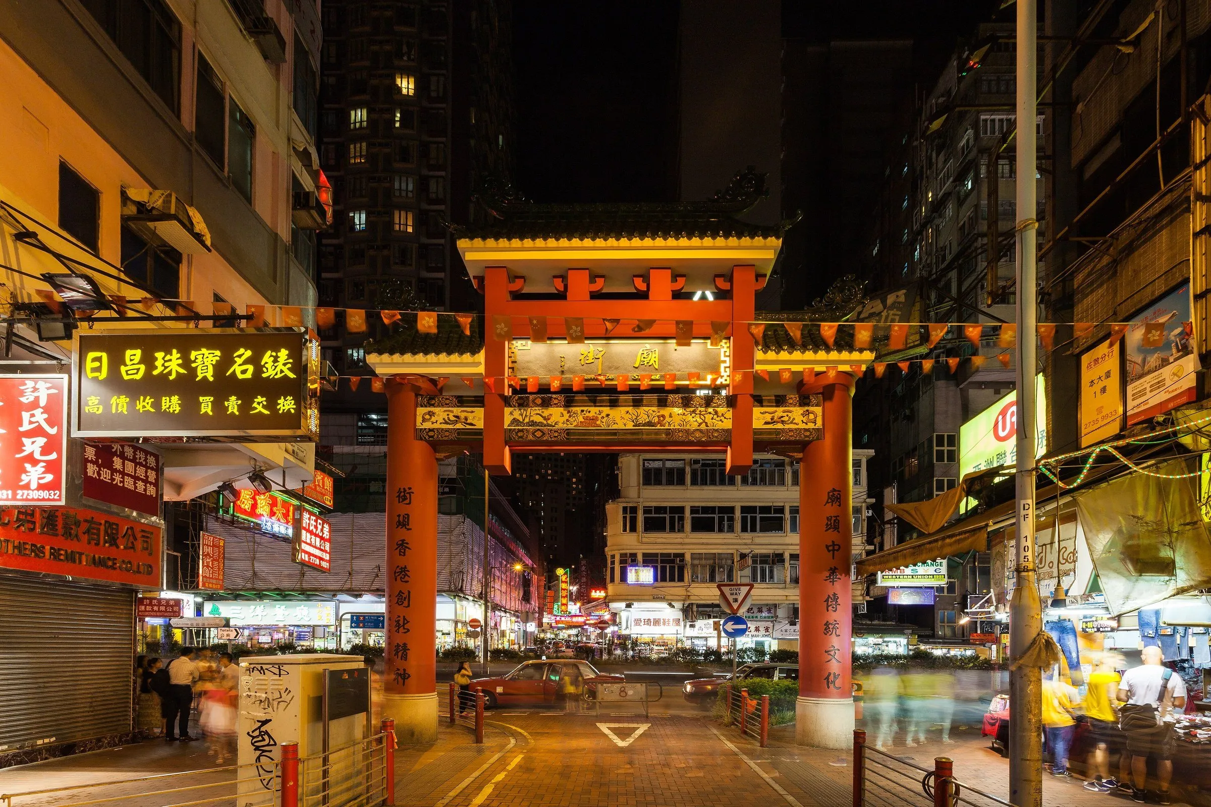 Pórtico vermelho da Temple Street, em Yau Ma Tei, à noite, com letreiros luminosos e a rua quase vazia ao fundo.