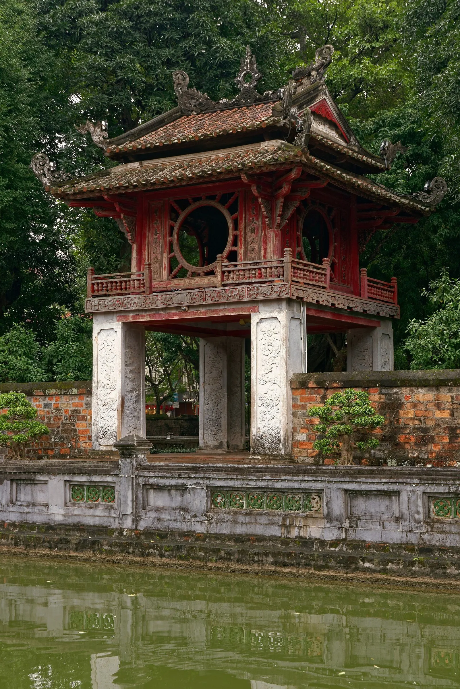 Pavilhão Khue Van de madeira vermelha no Templo da Literatura, com árvores e reflexo na água.