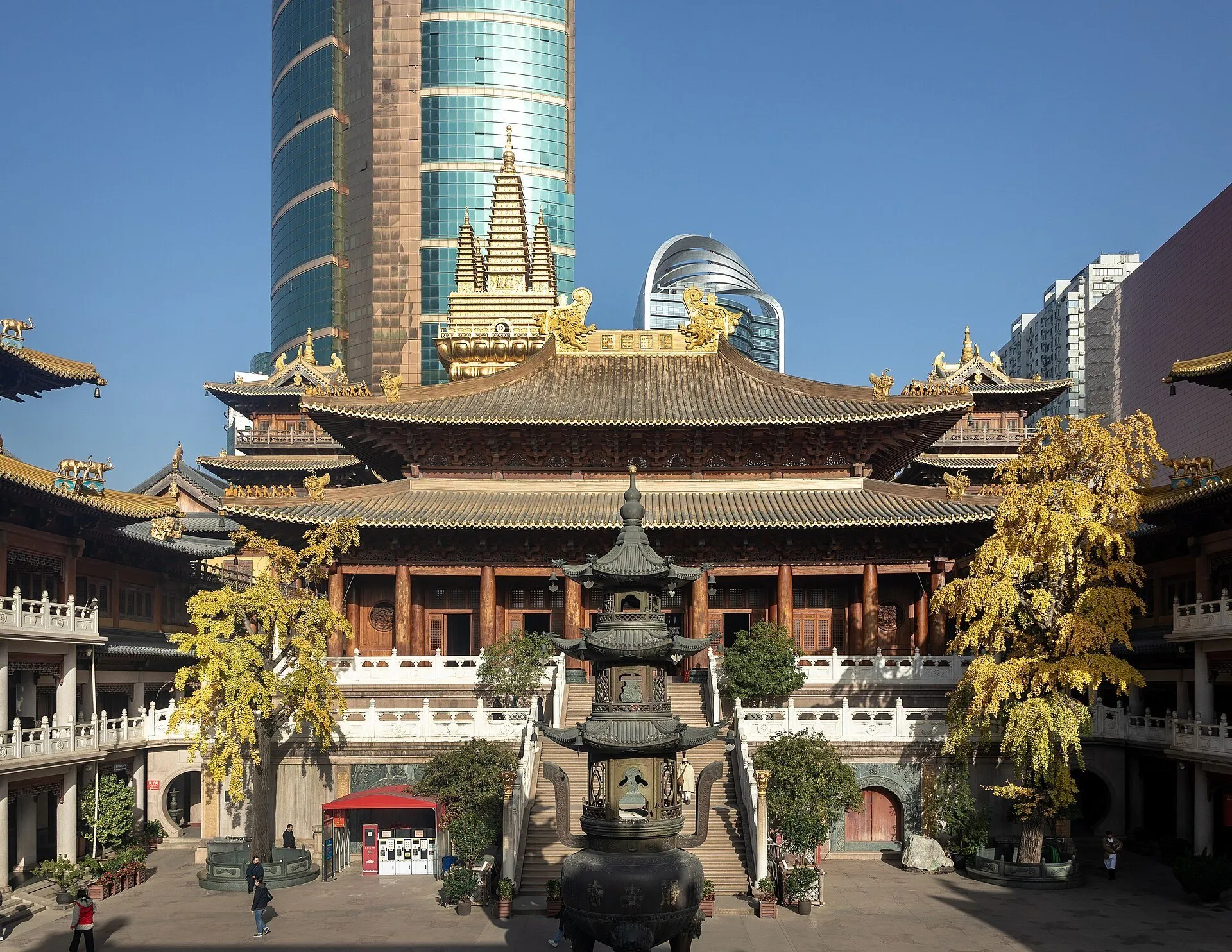 Courtyard of Jing’an Temple with an incense burner, yellow-leaved trees and golden roofs before tall buildings.