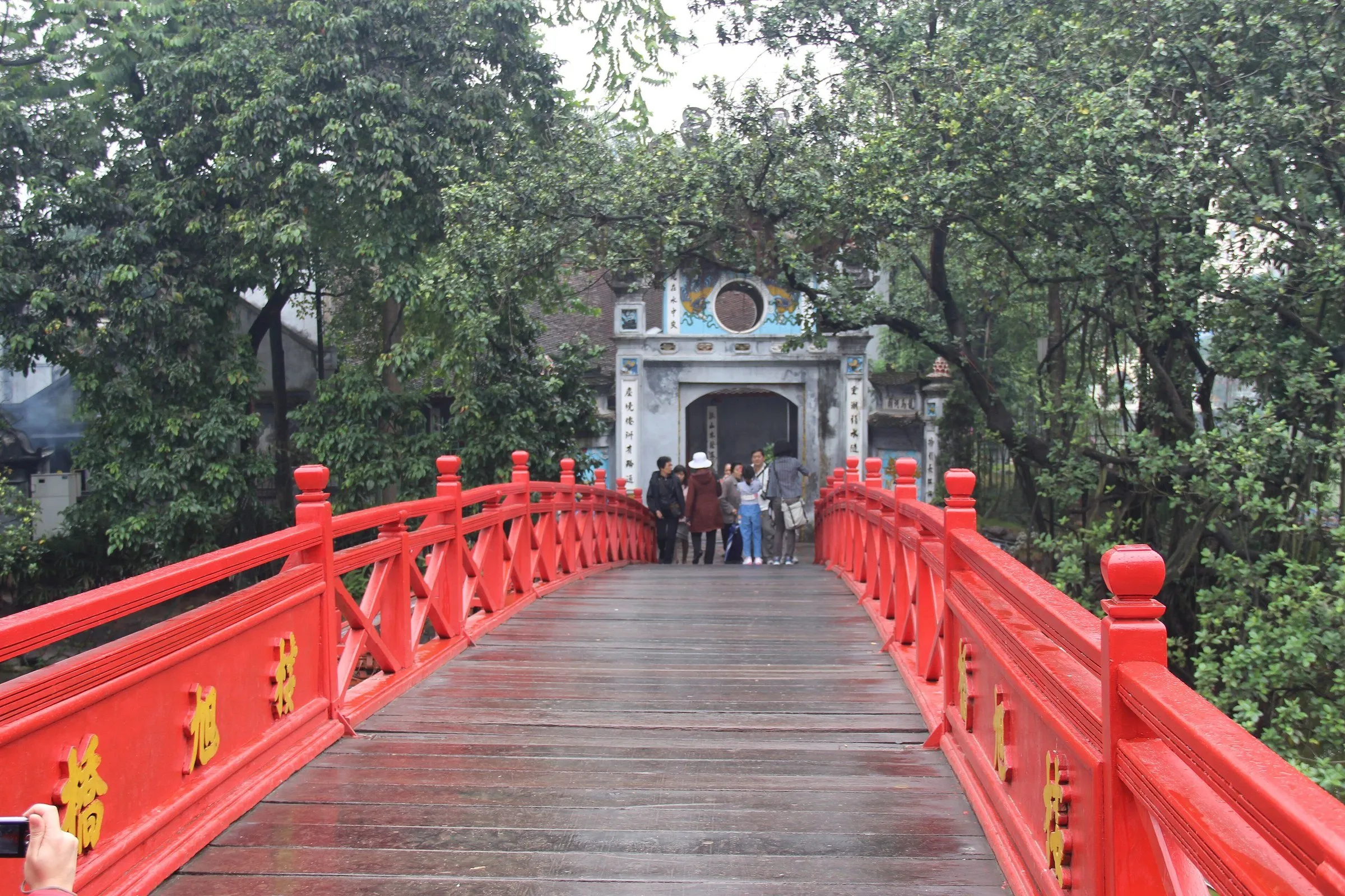 Ponte vermelha The Huc diante do portão do Templo Ngoc Son, entre árvores e visitantes.