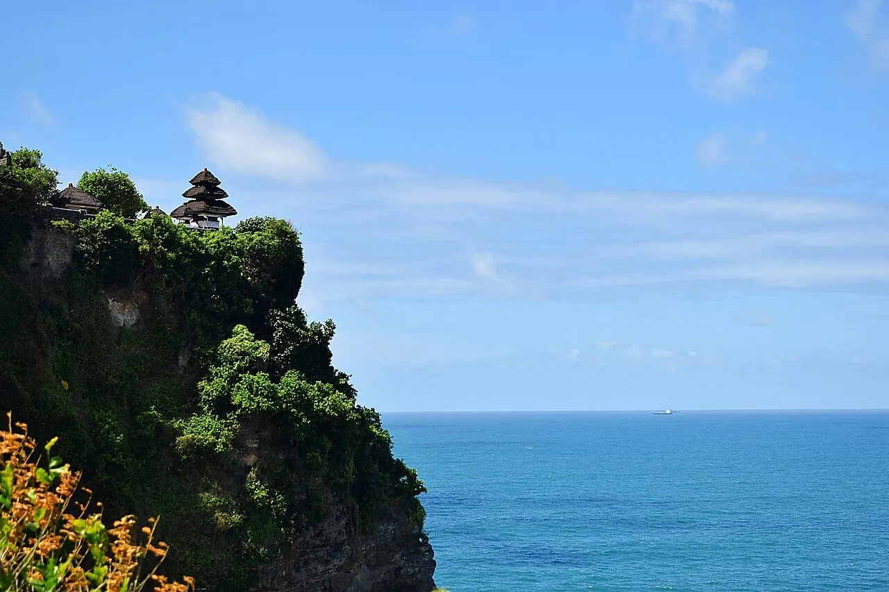 Templo de Uluwatu en lo alto de un acantilado cubierto de vegetación, frente al océano azul y un cielo claro.