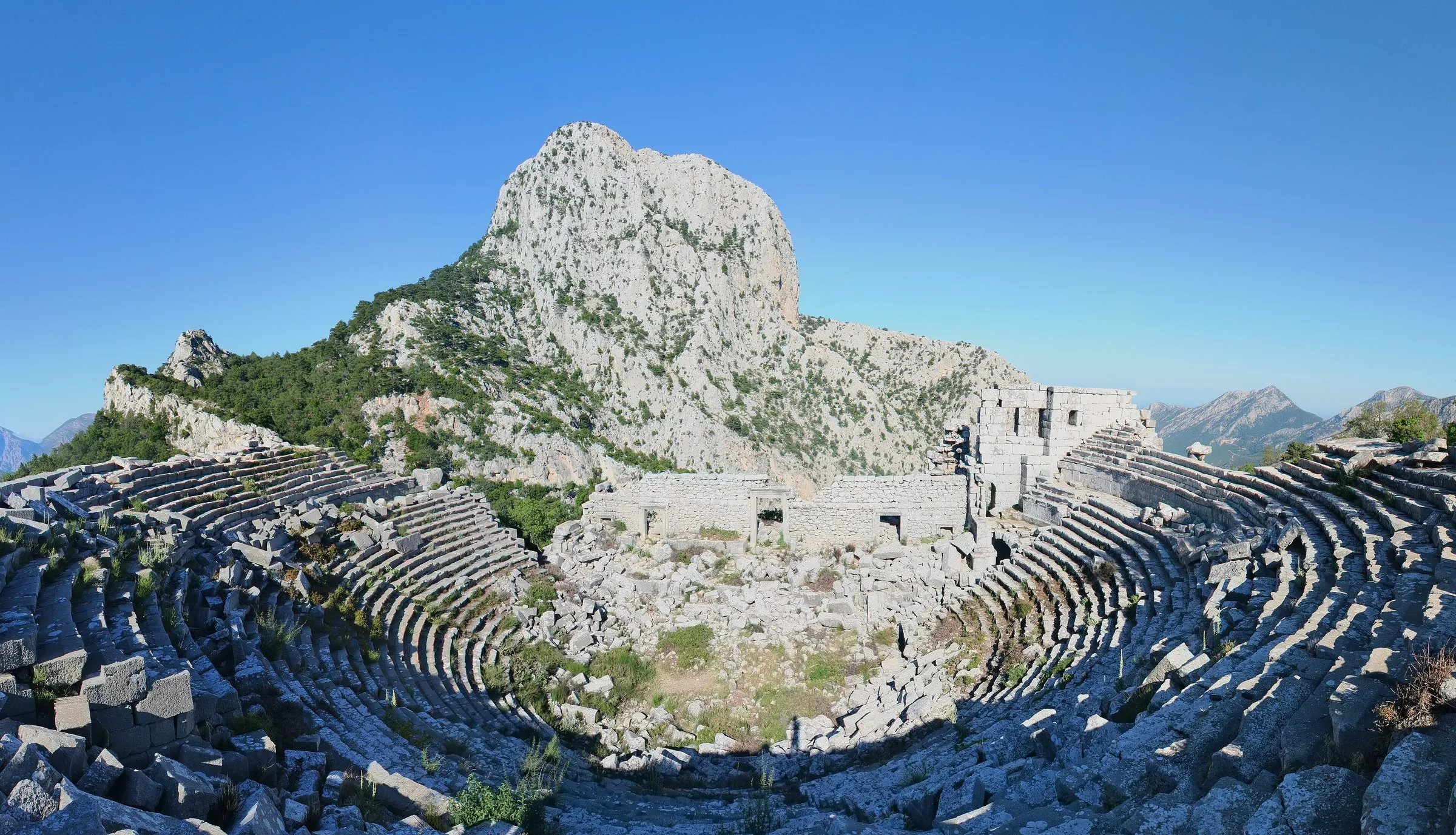 Teatro antigo de Termessos em ruínas, com arquibancadas em arco, um paredão de pedra ao fundo e montanhas ao longe.