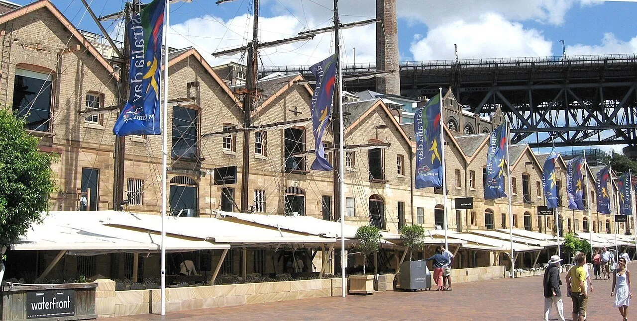 Stone facades in The Rocks, with flagpoles, flags, tables under awnings and the Harbour Bridge in the background.