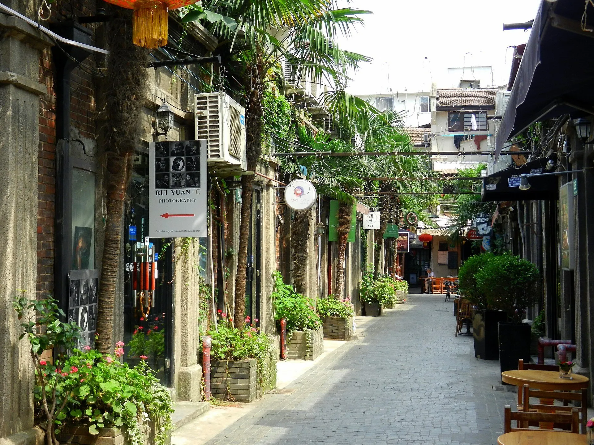 Paved Tianzifang alley with stone doorways, shops, potted plants and tables along the facades.