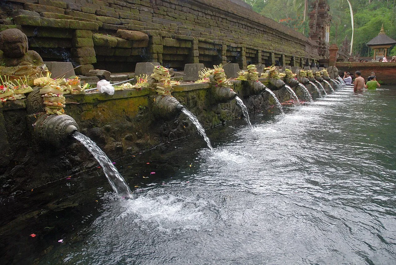 Hilera de caños de piedra vertiendo agua en una piscina de Tirta Empul, con ofrendas y fieles al fondo.