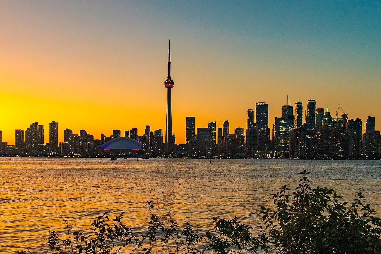 Torre CN e prédios de Toronto ao pôr do sol, com Lago Ontário e vegetação escura em primeiro plano.