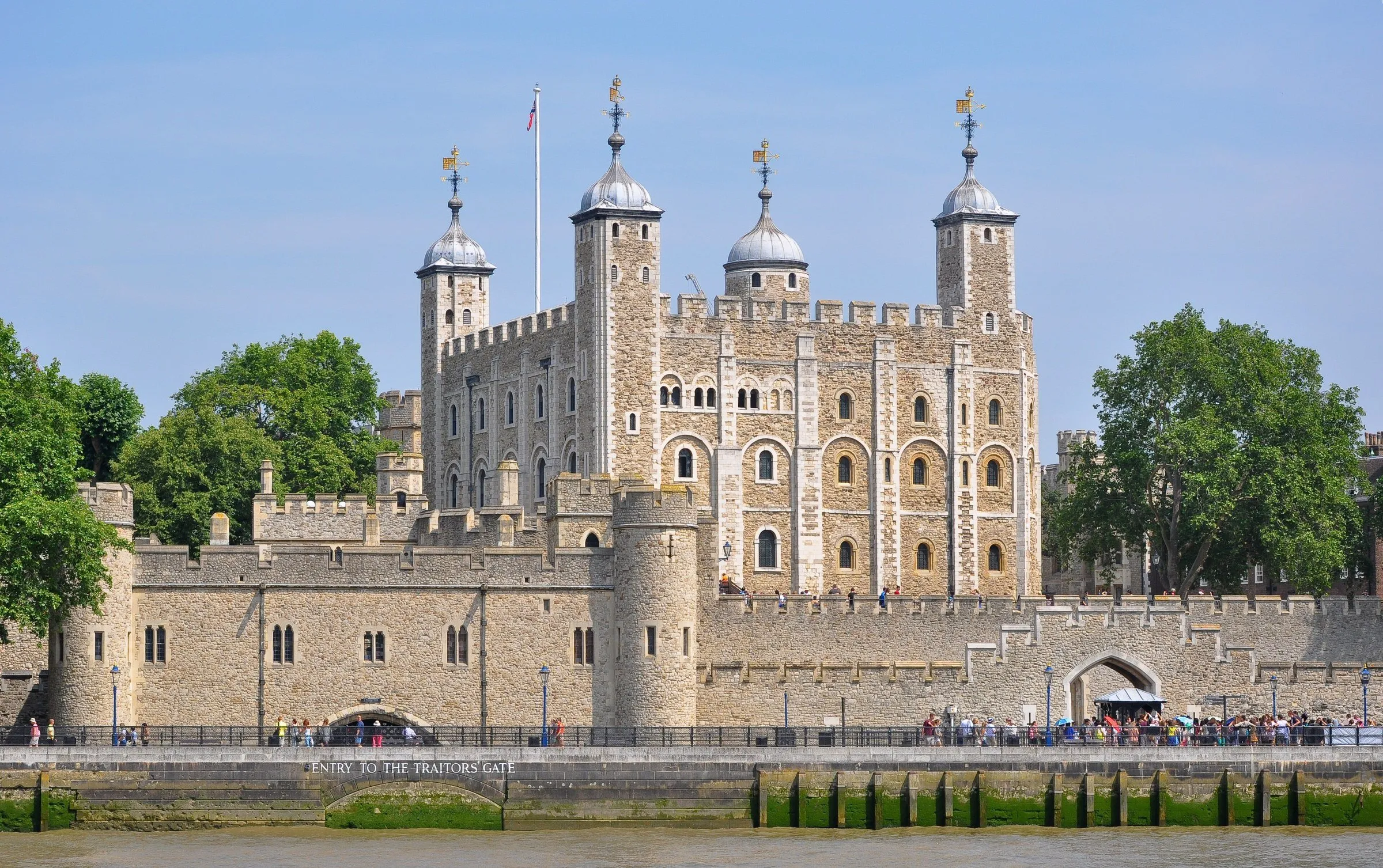 White Tower and stone walls of the Tower of London, seen from the Thames bank under a blue sky.