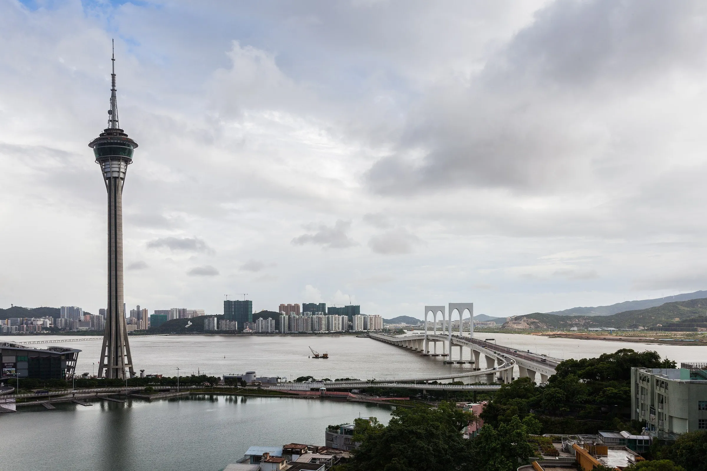 Torre de Macao vista desde el otro lado del agua, con la bahía, un puente en arco y la ciudad al fondo bajo un cielo nublado.