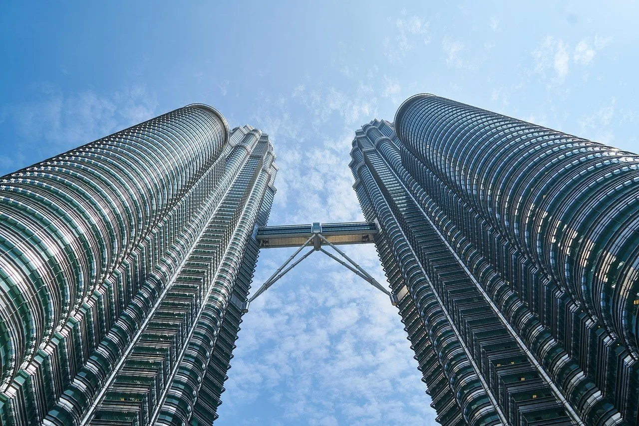 Vista de baixo para cima das duas Torres Petronas, com a passarela que as liga, contra um céu azul com nuvens finas.