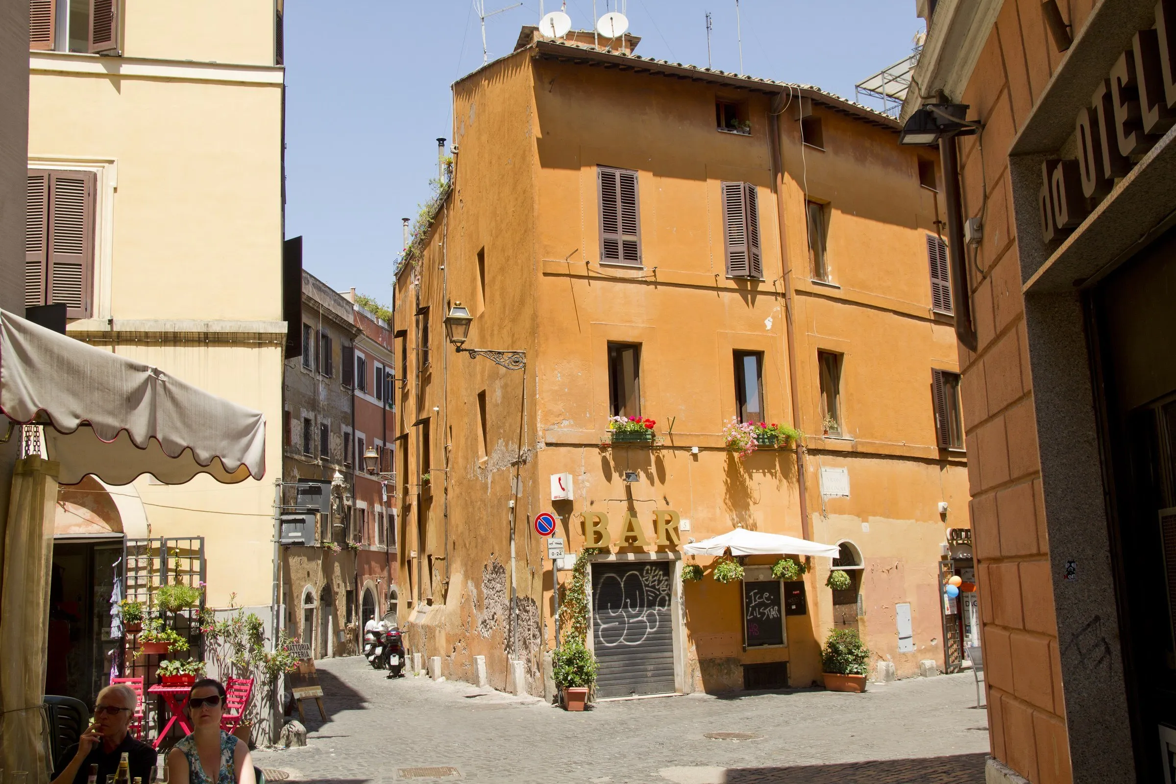 Trastevere corner with an ochre building, shuttered windows, bar tables on the sidewalk and a narrow stone street.