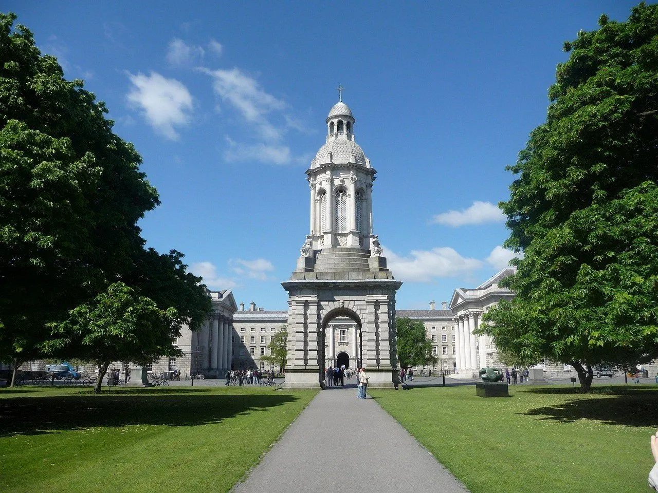 Campanario de piedra del Trinity College entre árboles, césped y edificios del campus, bajo cielo azul.