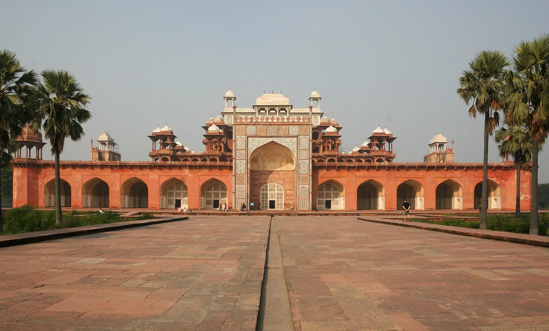 Fachada de la Tumba de Akbar en Sikandra, con patio de arenisca y palmeras a los lados.