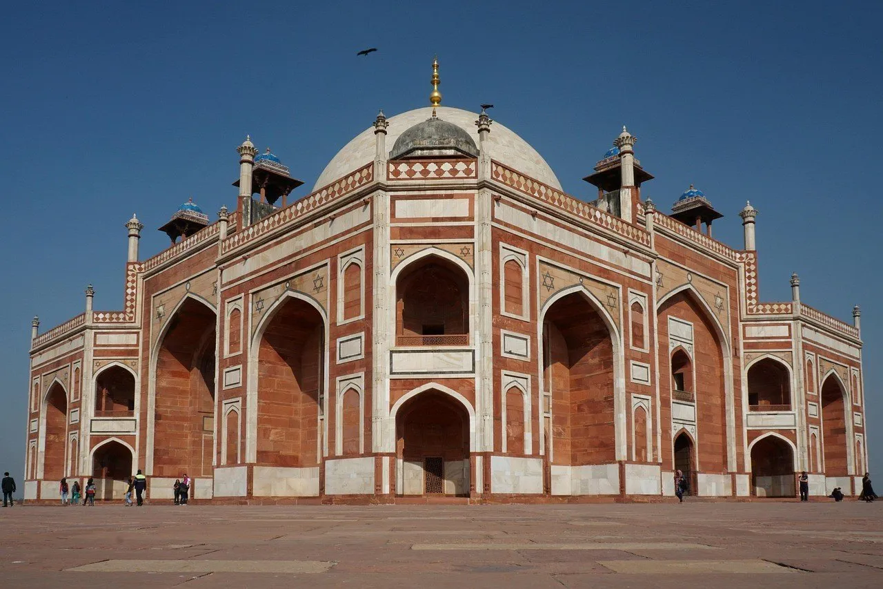 Fachada de la Tumba de Humayun, en Delhi, con arcos de arenisca roja, frisos blancos y cúpula blanca bajo cielo azul.