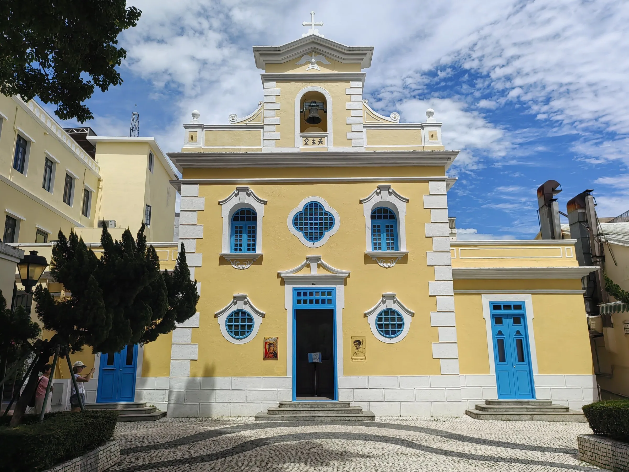 Fachada amarilla y blanca de la Capilla de San Francisco Javier, con puertas azules, en una plaza de empedrado ondulado.
