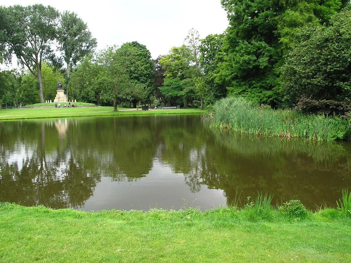 Lago do Vondelpark cercado de árvores e gramado, com monumento ao fundo.
