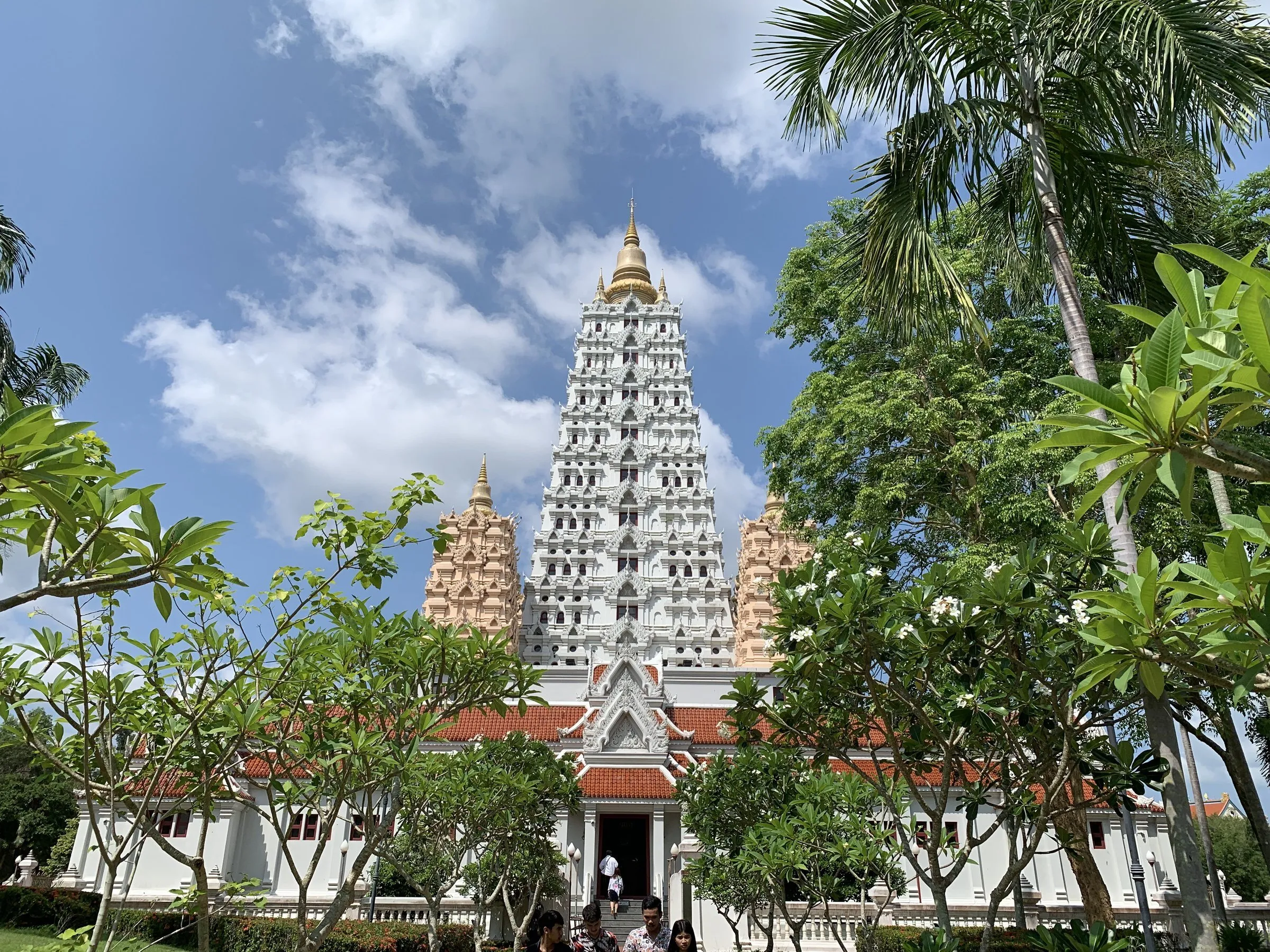 Torre branca e dourada do Wat Yansangwararam, com telhados vermelhos, árvores e visitantes na base.
