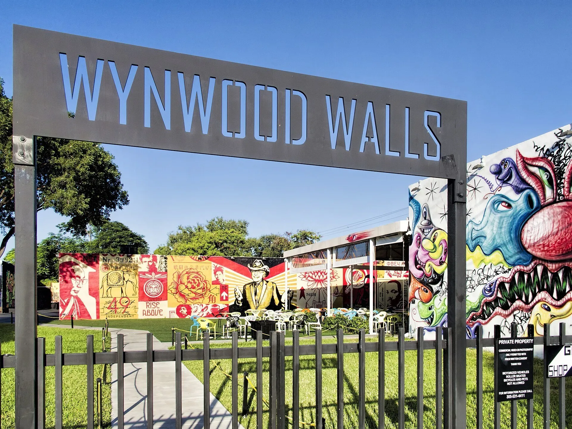 Letreiro de entrada do Wynwood Walls com portão, gramado, mesas e murais coloridos ao fundo.